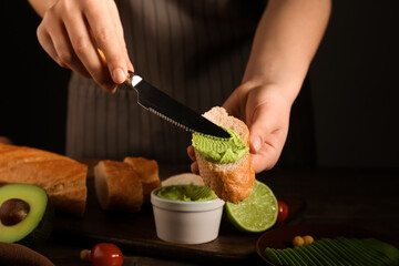Woman spreading tasty avocado hummus onto piece of bread at wooden table, closeup