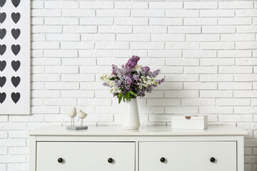 Vase with lilac flowers and decor on chest of drawers near white brick wall