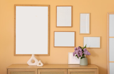 Chest of drawers with lilac flowers and blank frames on beige wall in room