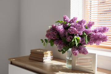 Vase with lilac flowers, books and frame on chest of drawers near window in room