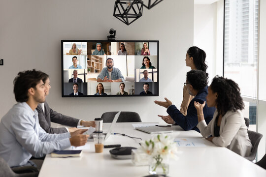 Team of businesspeople professionals participating in video conference gathered together in modern meeting room, on large screen displaying diverse colleagues actively engaged in virtual business call
