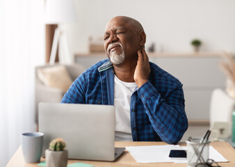 Naklejka premium Neckache. Senior African American Man Touching Aching Neck Suffering From Pain Sitting Working On Laptop In Modern Office. Healthcare, Health Problems In Older Age Concept