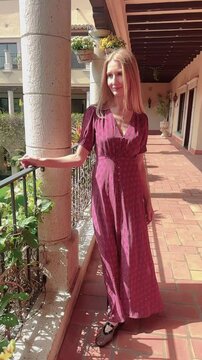 Vertical video shows a young woman with long blonde hair, dressed in a long, red dress, walking in slow motion on the terrace of a Spanish-style villa.  