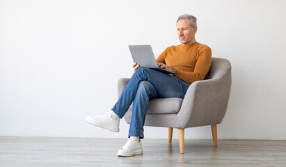 Portrait of focused mature man using laptop sitting on armchair. Confident male adult working on...