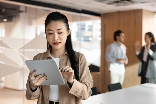 Focused Vietnamese businesswoman working on digital tablet device standing in meeting room, colleagues in background. Professional workflow using modern innovative business AI tool, apps and platforms