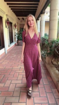 Vertical video shows a young woman with long blonde hair, dressed in a long, red dress, slowly walking towards the camera on the terrace of a Spanish-style villa.  