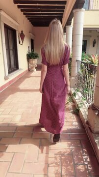 Vertical video shows a young woman with long blonde hair, dressed in a long, red dress, slowly walking, facing backwards, on the terrace of a Spanish-style villa.  