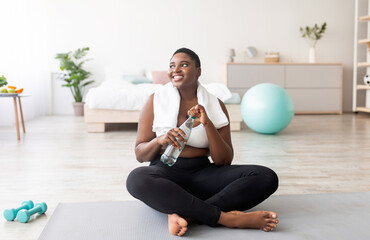 Plump black lady in sportswear sitting on sports mat with bottle of clear water at home, full...