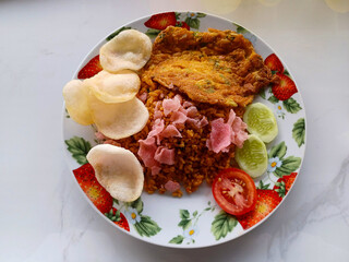 A vibrant plate of Nasi Goreng, a popular Indonesian fried rice dish. The rice, a rich brown hue, is topped with a golden-yellow omelet, crispy shrimp crackers, sliced cucumbers and tomatoes