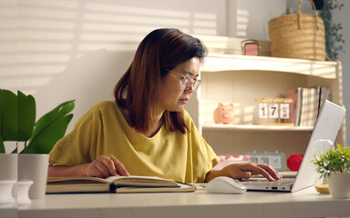 Asian woman works from home, surrounded by notes, a laptop, and messages. She browses the internet, writes ideas, and researches for her startup, utilizing wireless technology.