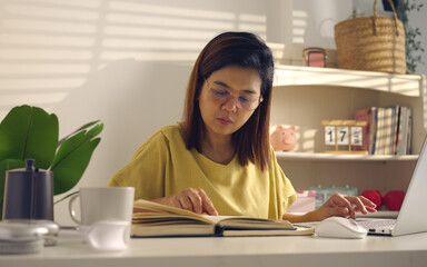Asian woman works from home, surrounded by notes, a laptop, and messages. She browses the internet, writes ideas, and researches for her startup, utilizing wireless technology.