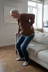 Ill senior man clutches painful spine unable to stand from couch due to multiple health issues, suffering from hernia. Herniated disc agony, pinched nerves, chronic kidney disease, mobility loss. 