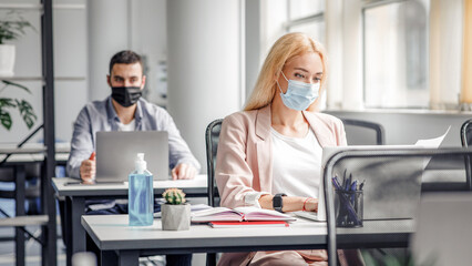 New working conditions and social distancing after quarantine. Blonde woman in protective mask works with documents and laptop at workplace with notepad and big bottle of antiseptic in interior