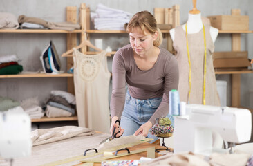 Calm and attentive young female dressmaker working in tailor artisan workshop, using fabric shears...