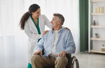 Obraz premium Portrait of happy senior man in wheelchair and his female nurse at retirement home. Mature male patient with disability and his caregiver posing and smiling, looking at each other indoors
