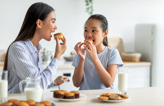 Having a bite. Cute asian girl and her mother eating snacks in kitchen, happy woman and her adorable female child enjoying homemade muffins and drinking milk together