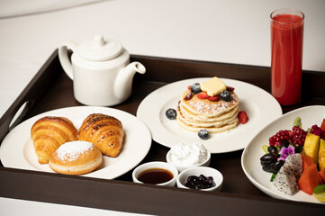 Close-up of a breakfast-in-bed tray with coffee kettle, pancakes, croissant, fruit salad, watermelon juice, jams, and ice cream on white bedsheets in soft natural and artificial light.