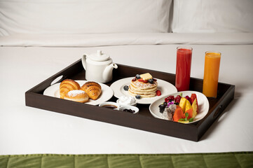 Breakfast-in-bed tray with coffee kettle, pancakes, croissant, fruit salad, orange juice, watermelon juice, jams, and ice cream on white bedsheet in soft natural light.