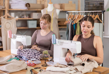 Two young female seamstresses working side by side at sewing machines in tailoring studio, focusing on assembling fabric pieces with precision and control