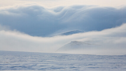 Winter Arctic landscape. View of snow-covered tundra. Mountains shrouded in clouds are visible in the distance. Low clouds and fog. Harsh polar climate of the northern regions of Russia. Chukotka.