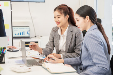 Two Asian businesswomen sit together working, using a laptop to check documents.