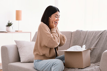 Side view of excited asian female buyer received good parcel, looking at cardboard box at home, satisfied with great purchase, happy lady unpacking package, overjoyed by postal shipping delivery
