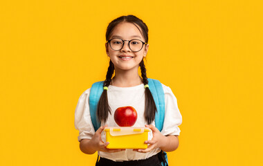 School Nutrition. Smiling Chinese Schoolgirl Holding Lunch Box Posing On Yellow Background. Studio Shot