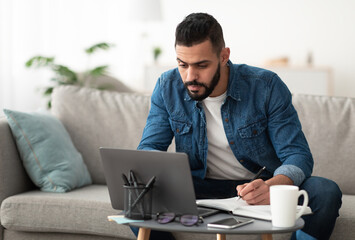 Serious young Arab man using laptop, working remotely, taking notes during remote business meeting from home. Millennial Eastern guy studying online, writing down info. Modern technologies concept