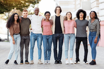 Friendship, community, team concept. Multiracial group of happy millennial men and women posing on the street, standing in a row, embracing and cheerfully smiling at camera, full length shot