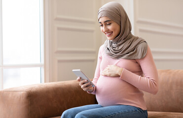 Happy Pregnant Muslim Woman Using Smartphone Texting And Browsing Internet On Phone Sitting On Sofa...