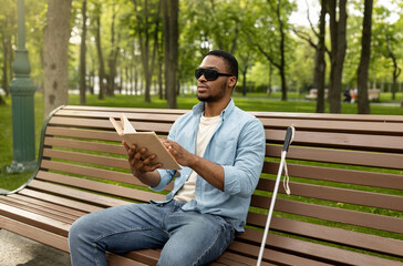 Obraz premium Young black visually impaired man sitting on bench in city park, reading Braille book outdoors. Millennial blind guy in dark glasses studying or enjoying entertainment literature, outside