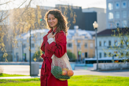 Young woman in a red coat carrying a reusable mesh bag filled with fresh produce, strolling through a vibrant city park while embracing eco-conscious choices