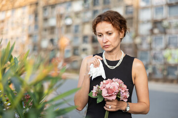 Fototapeta premium Upset woman in black dress, who lost loved one, brings flowers to a grave