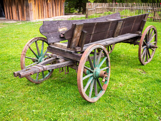 Old wooden wagon with rusty iron parts standing on green grass near a wooden building. A...