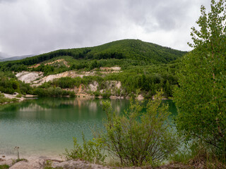 Panoramic view of a flooded quarry with green water and rocky terraces, surrounded by forested hills. Reflections of cliffs and sky visible on the calm surface. Hiking spot near Kraľovany.