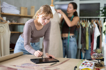Attentive dressmaker drawing dress design in tablet while other working with mannequin in sewing workshop