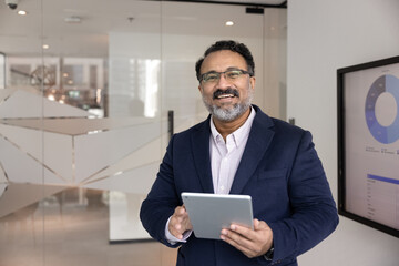 Portrait of happy Indian male executive or CEO dressed in formal suit stands in modern office,...