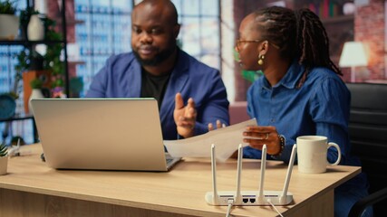 Fototapeta premium Black irritated man and woman rebooting their wireless router and checking network settings, feeling angry with the struggles to resolve an internet outage disrupting their tasks. Camera A.