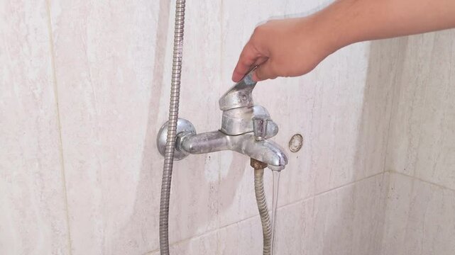 A close-up video showing a person&rsquo;s hand lowering the lever to turn off an old metal faucet with a flexible shower hose, set against a tiled bathroom wall.