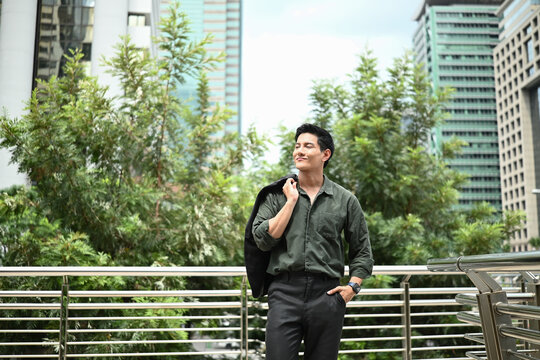 A man wearing a green shirt and black jacket stands on a bridge in front of a city skyline.