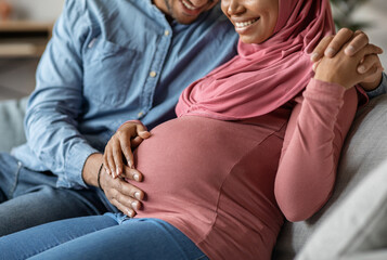 Happy Pregnancy. Loving Husband Tenderly Touching Belly Of His Pregnant Muslim Wife While They Sitting Together On Couch In Living Room, Unrecognizable Islamic Spouses Enjoying Upcoming Parenthood