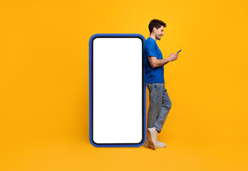 Great App. Happy young guy using cell standing leaning on big white smartphone screen with blue case on yellow studio wall, full body length. Mock up for website app, free copy space for advertisement © Prostock-studio