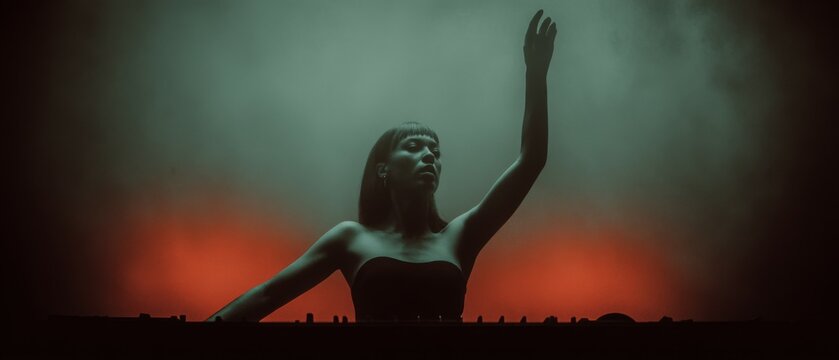A female DJ commands the stage, one hand raised triumphantly amid swirling fog. Warm rim lights trace her silhouette while lasers slice through the smoky background. A high-energy live DJ performance.