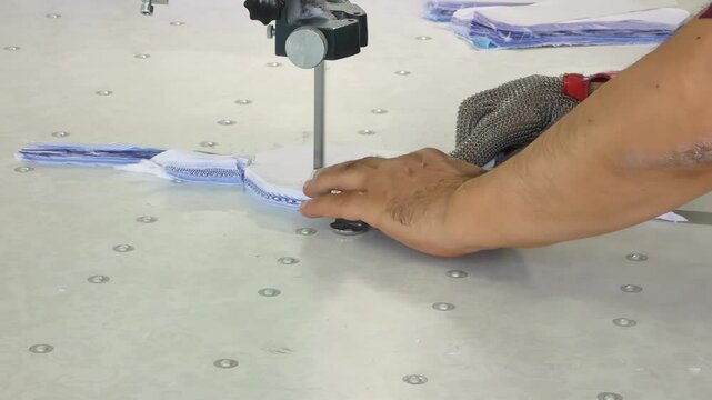 The commercial textiles worker works on the machine cutting a pile of fabric at the clothing factory