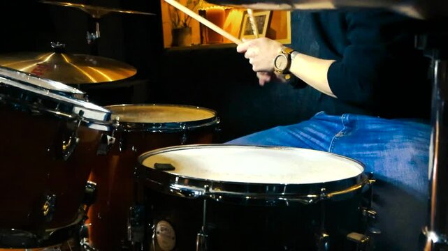 Lateral view of man playing drums in dimly lit studio room. Musician hands with drumsticks beating drums and strike the cymbals. Soft glow on drum set. Drummer beating rhythm, scenic drumming angles