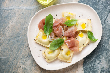 Plate of italian ravioli served with prosciutto and green basil, horizontal shot on a beige and grey granite background, high angle view