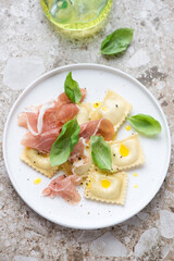 White plate with ravioli, prosciutto and green basil on a light-brown granite background, vertical shot, high angle view