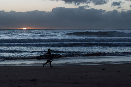 Surfer walks along Manly Beach at sunrise under moody clouds