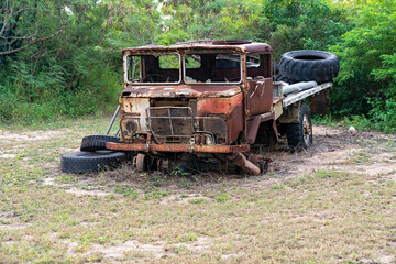 Rusty abandoned truck sitting on overgrown grass in front of thick green trees, missing windows and parts.