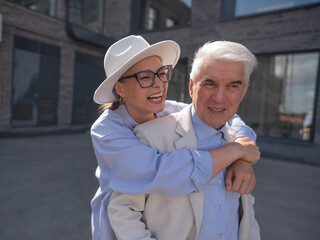 A woman in a hat and glasses hugs an elderly man in a white jacket from behind. Romantic relationships of mature people. 